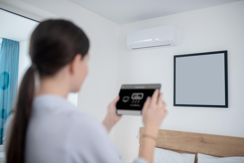 Kobieta trzyma tablet, na którym wyświetla się interfejs do sterowania inteligentnym domem. W tle, na białej ścianie w sypialni, widoczny jest klimatyzator, którym użytkowniczka prawdopodobnie zarządza zdalnie.